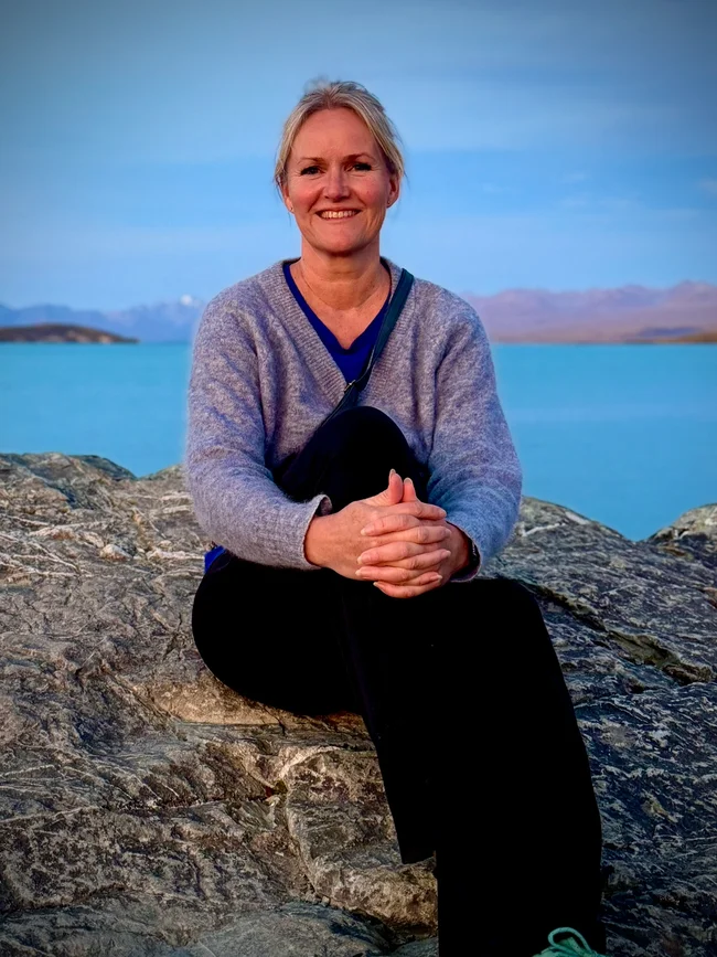 Lene sitter på en stein ved et turkisblått vann med fjell i bakgrunnen.
